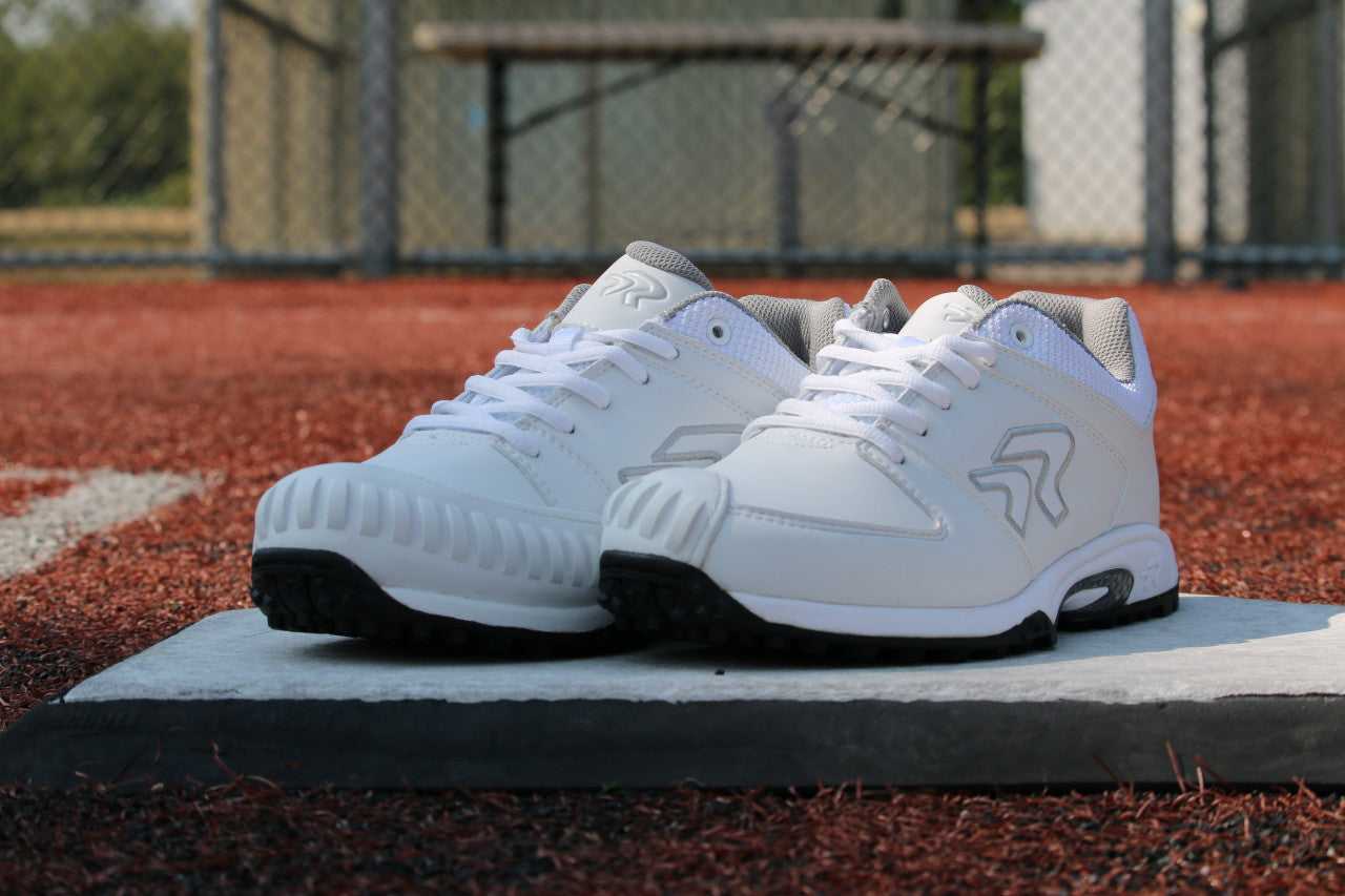 A pair of Ringor Flite Turf PTT Women's Fastpitch Softball Shoes with Pitching Toe (4842S) are displayed on a base atop red artificial turf, with gray accents, a metal fence, and bench in the background.