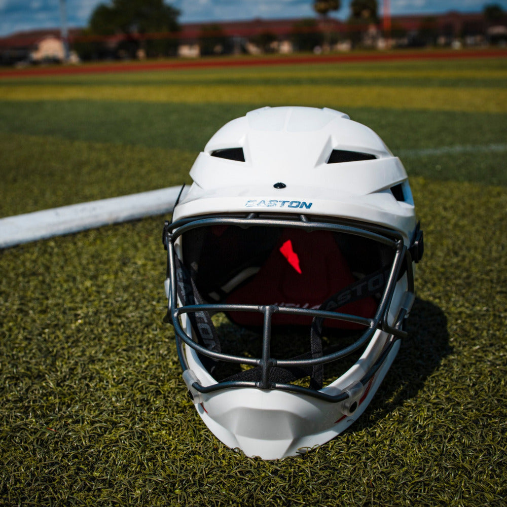 A white Easton Hellcat Slowpitch Fielding Helmet (EHCATH) with a black metal cage from Easton sits on green turf near a foul line, with a blurred field and trees in the background.