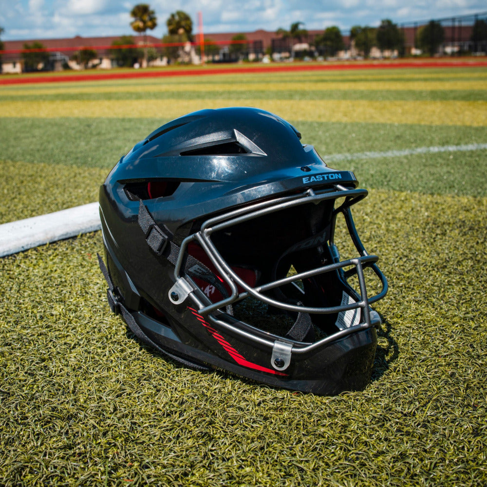 An Easton Hellcat Slowpitch Fielding Helmet (EHCATH) by Easton, featuring a black shell and silver face guard, rests on artificial turf near a foul line with a baseball field and school buildings under a blue sky in the background.