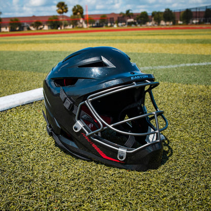 An Easton Hellcat Slowpitch Fielding Helmet (EHCATH) by Easton, featuring a black shell and silver face guard, rests on artificial turf near a foul line with a baseball field and school buildings under a blue sky in the background.