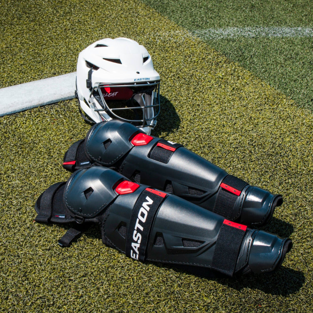 A white helmet and black Easton Hellcat Slowpitch Fielding Leg Guards (EHCATL) by Easton lie on green artificial turf near a white boundary line under bright sunlight.