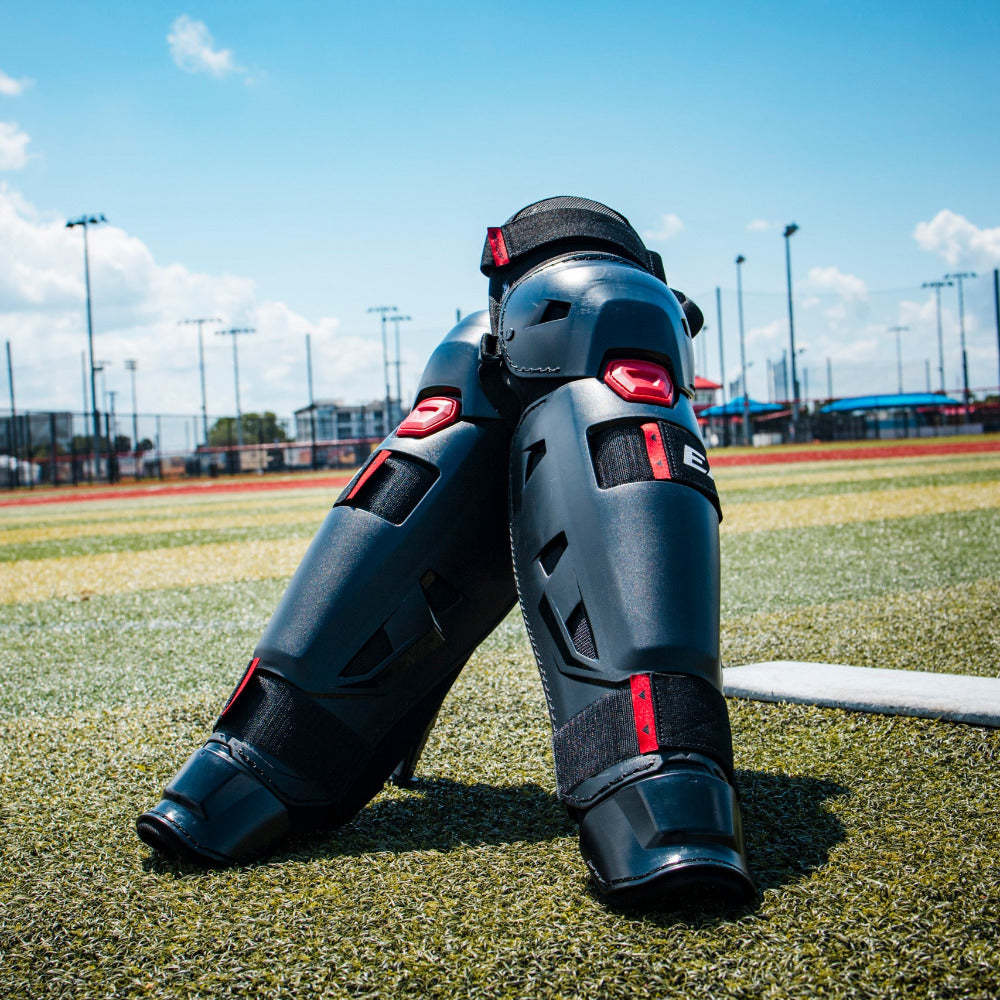 A close-up of Easton Hellcat Slowpitch Fielding Leg Guards (EHCATL) by Easton, in black and red, resting on an outdoor sports field with a blue sky, fences, buildings, and poles in the background.