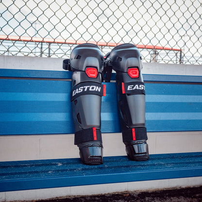A pair of Easton Hellcat Slowpitch Fielding Leg Guards (EHCATL) in black and red rest upright against blue stadium seats, with a chain-link fence behind—ideal protection for slowpitch softball on game day.