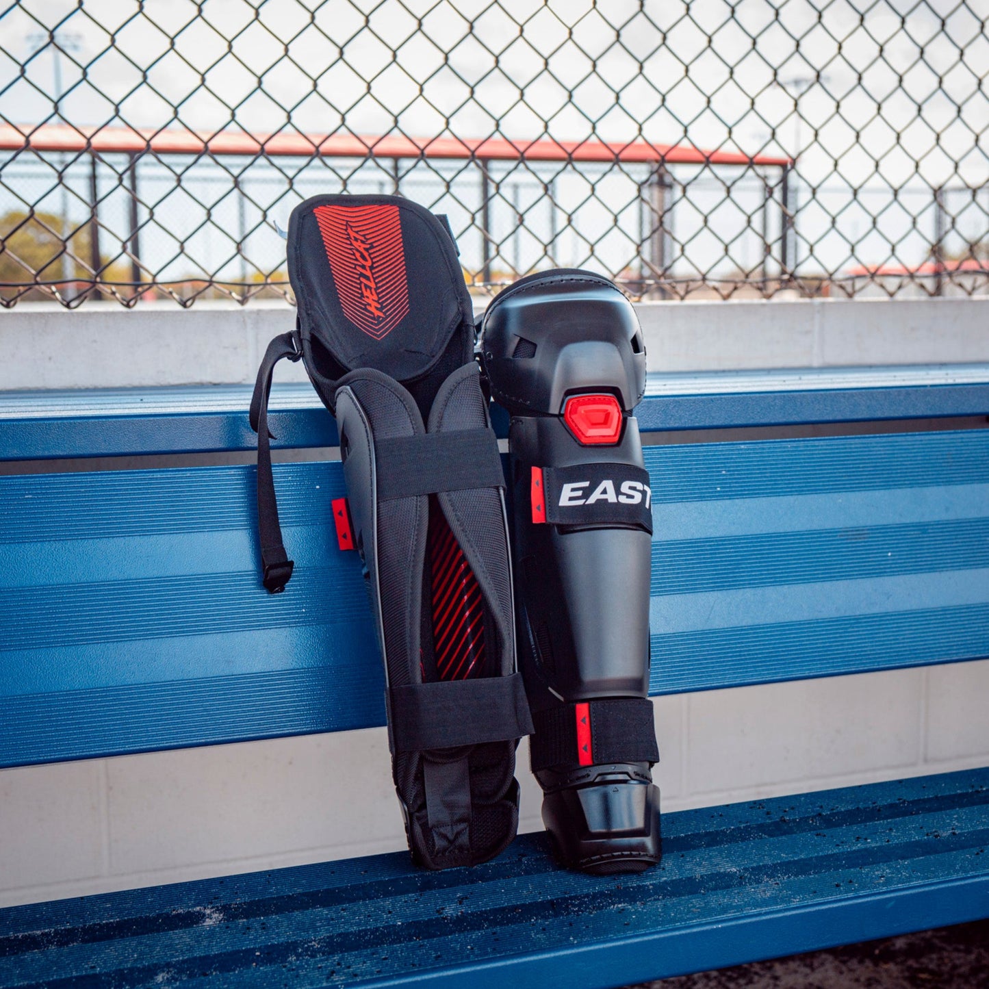 A pair of black and red Easton Hellcat Slowpitch Fielding Leg Guards (EHCATL) with the Easton brand name leans against a blue bench near a chain-link fence at an outdoor slowpitch softball field.