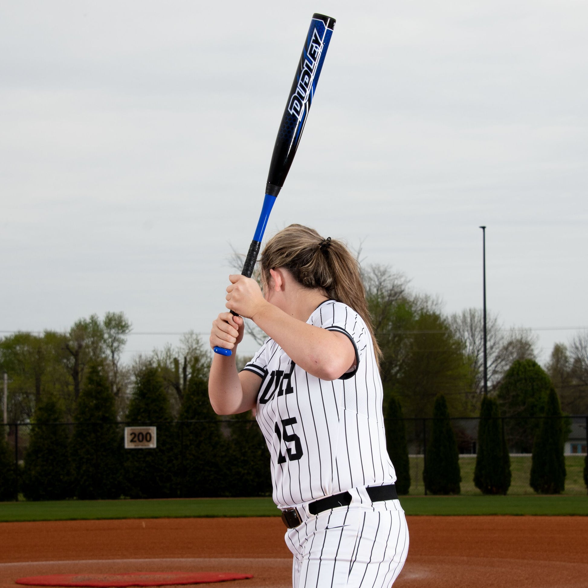A softball player in a white and black pinstripe uniform gets ready to bat on the field, holding her 2023 Dudley Doom (-10) Fastpitch Softball Bat: DDFP10 by Dudley. The sky is overcast, with trees visible in the background.