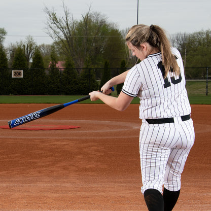 A softball player in a white pinstripe uniform with #19 and a blonde ponytail swings the 2023 Dudley Doom (-10) Fastpitch Softball Bat: DDFP10 by Dudley on a dirt field, with trees and a fence in the background, viewed from behind.
