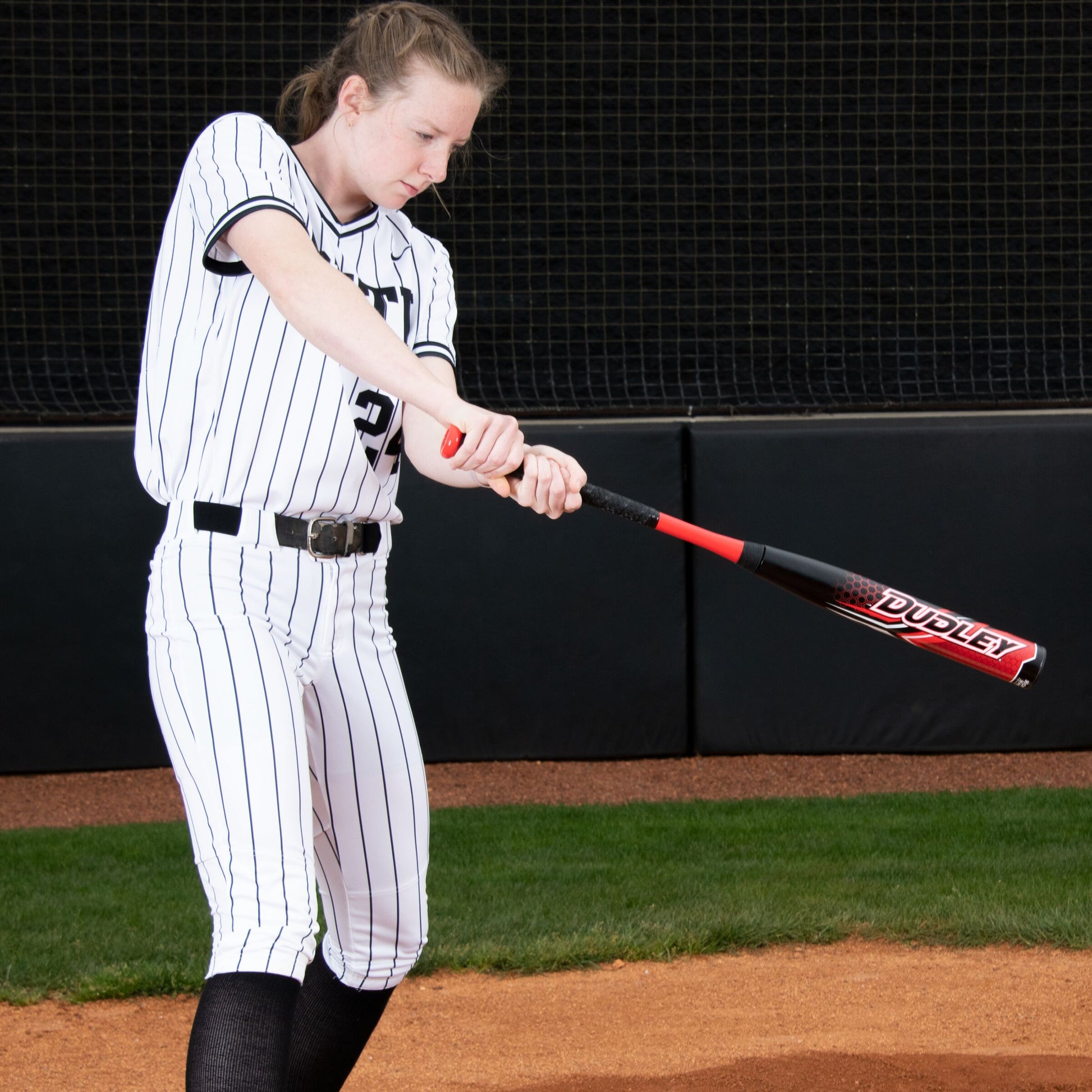 A softball player in a white pinstriped uniform swings the Dudley 2023 Doom (-11) Fastpitch Softball Bat (DDFP11) on a field with dirt, green grass, and a black fence in the background.