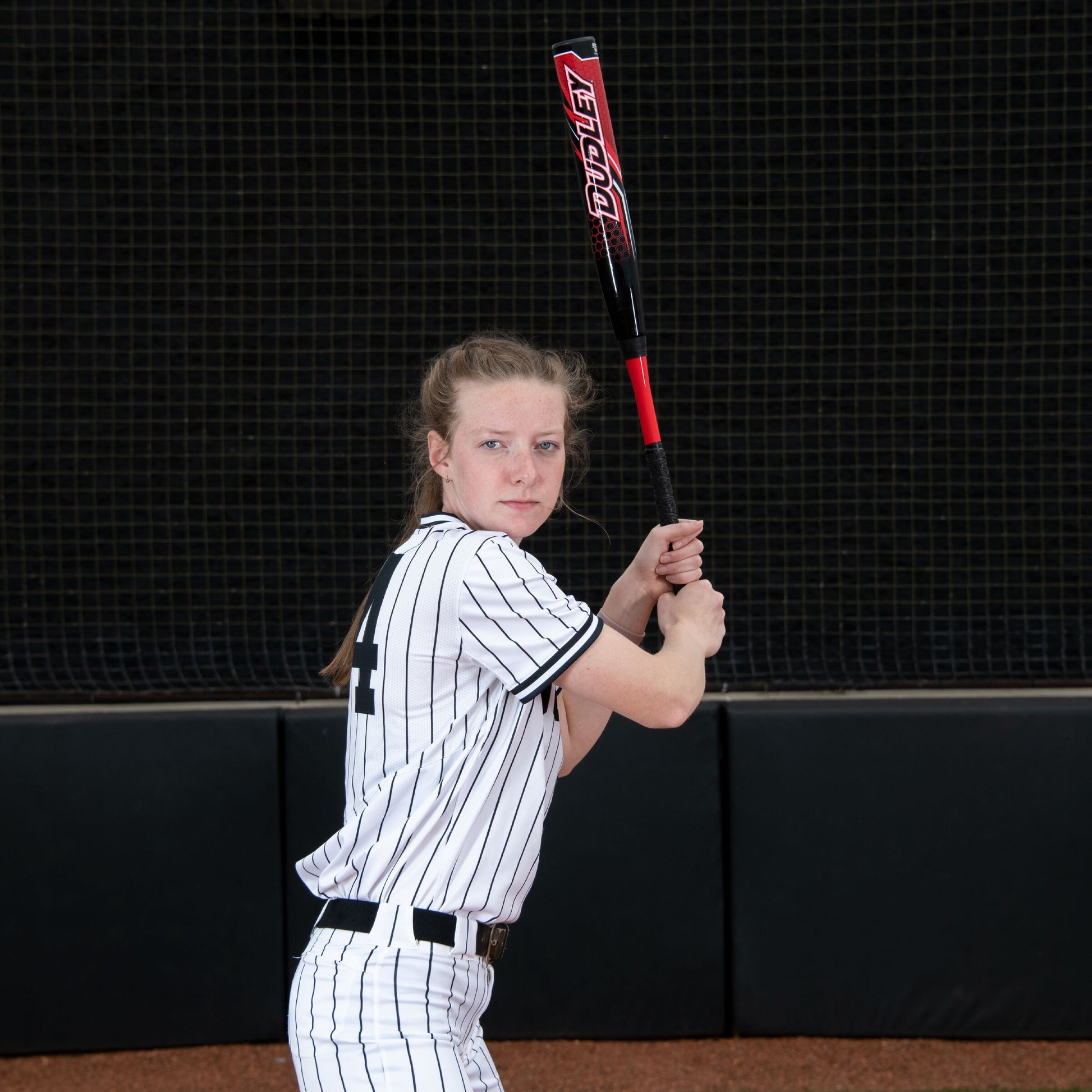 A young female player in a white pinstripe uniform grips the 2023 Dudley Doom (-11) Fastpitch Softball Bat (DDFP11) by Dudley, ready to swing on a field with a dark netted background.