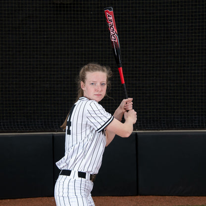 A young female player in a white pinstripe uniform grips the 2023 Dudley Doom (-11) Fastpitch Softball Bat (DDFP11) by Dudley, ready to swing on a field with a dark netted background.
