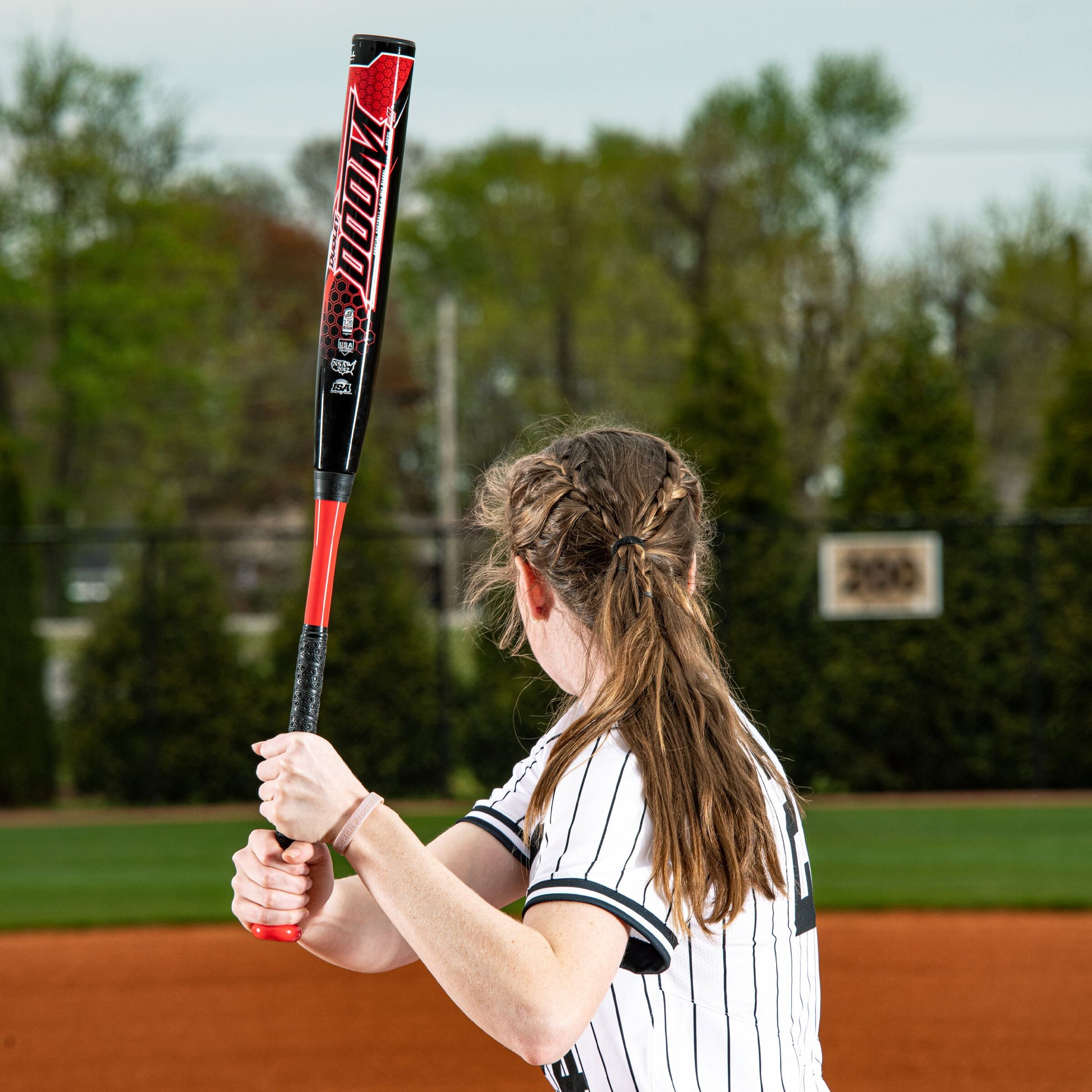 A softball player with braided hair stands ready to bat, gripping a 2023 Dudley Doom (-11) Fastpitch Softball Bat by Dudley on a field featuring trees and a scoreboard in the background.