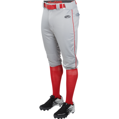 A person is wearing Rawlings Adult Launch Knicker Baseball Pants with Piping (Blue Grey/Scarlet), featuring red piping, a red belt, red knee-high socks, and black baseball cleats. The top half of the person is not visible.