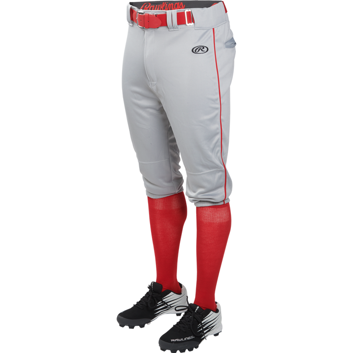 A person is wearing Rawlings Adult Launch Knicker Baseball Pants with Piping (Blue Grey/Scarlet), featuring red piping, a red belt, red knee-high socks, and black baseball cleats. The top half of the person is not visible.