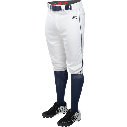 A person is shown from the waist down wearing Rawlings Adult Launch Knicker Baseball Pants with Navy piping in white/navy, a black Rawlings belt, navy socks, and black Rawlings cleats against a white background.