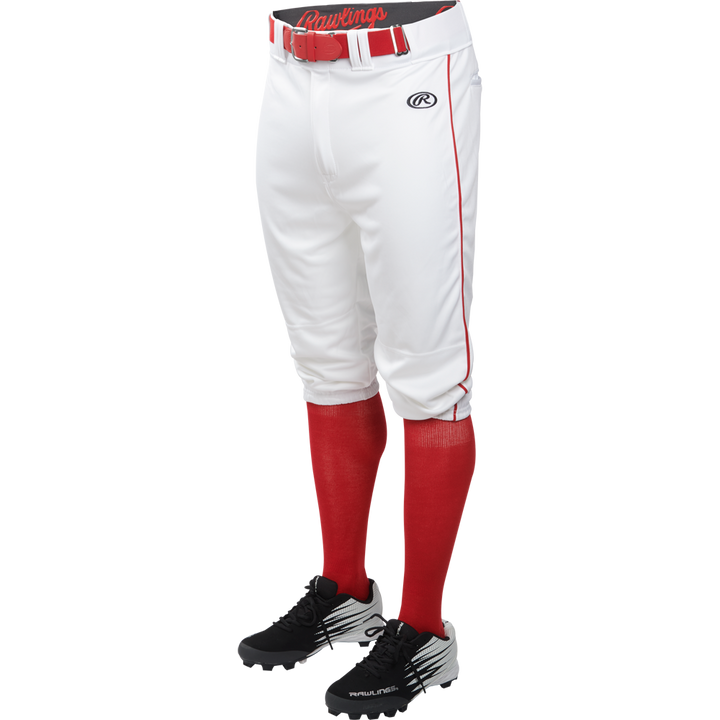 A person in Rawlings Adult Launch Knicker Baseball Pants with white/scarlet piping, a red Rawlings belt, red socks, and black cleats stands against a plain white background.