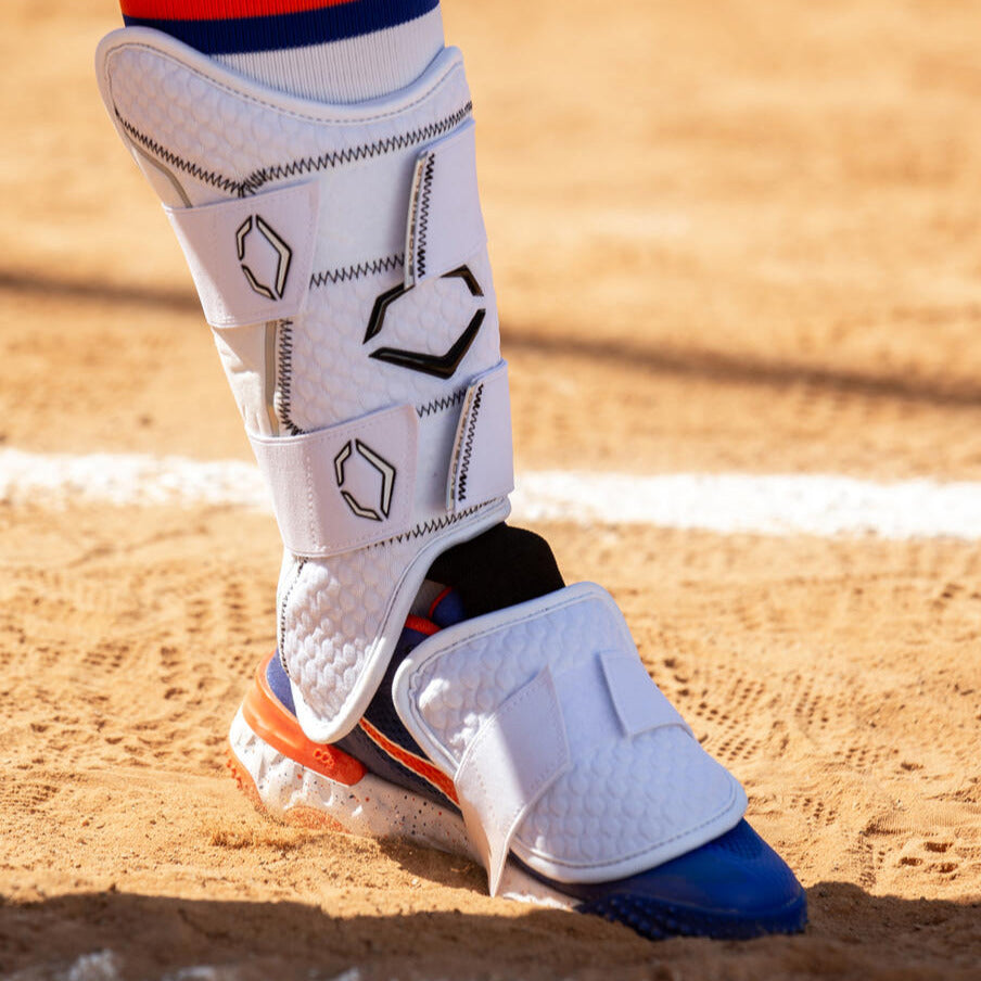 A close-up of the EvoShield PRO-SRZ 2.0 Batter's Leg Guard (WB5726901) protecting a player’s leg over blue, orange, and white cleats on a sandy field by a crisp white chalk line.