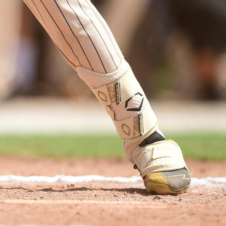 A close-up of a baseball player's lower leg and foot wearing an EvoShield PRO-SRZ 2.0 Batter's Leg Guard (WB5726901) and cleats, stepping onto the dirt near the foul line on a baseball field.