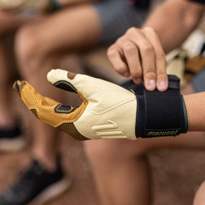 A person wears Marucci Blacksmith Adult Batting Gloves (MBGBKSMFW) in beige and brown with a durable goatskin palm, while blurred people sit in the background.