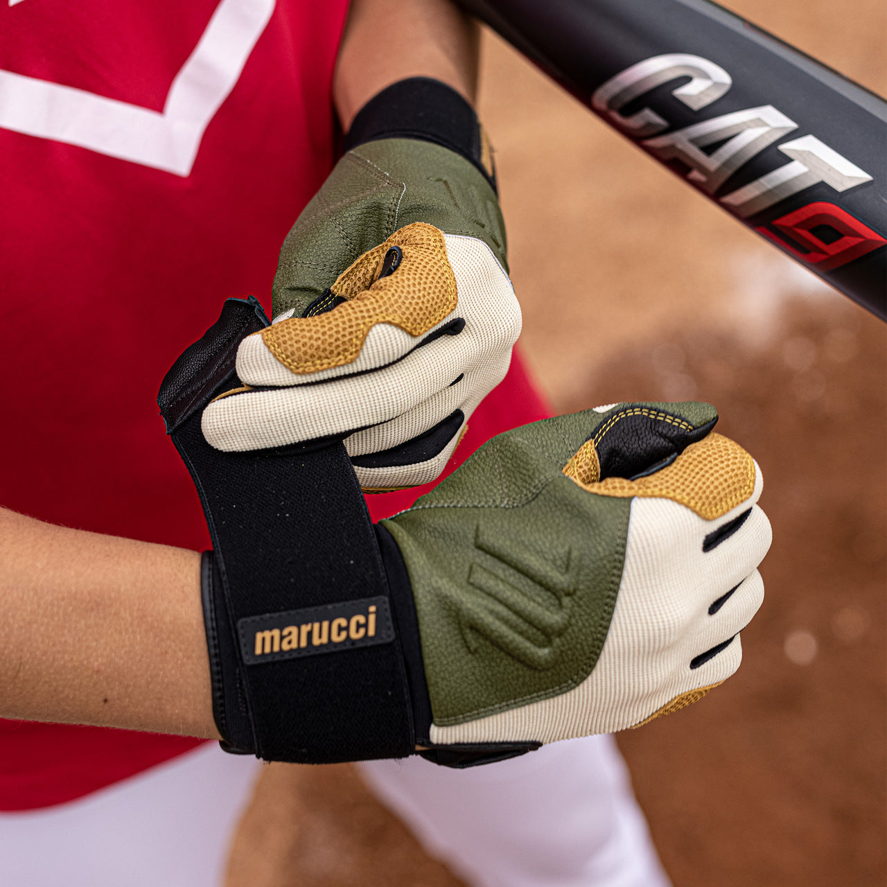 A person wearing green, yellow, and white Marucci Blacksmith Adult Batting Gloves (MBGBKSMFW) fastens one glove's strap while holding a Marucci CAT9 baseball bat on a dirt field.