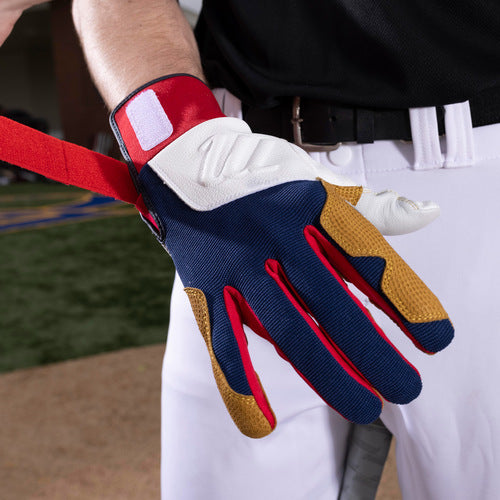 A person in a black shirt and white pants fastens the strap of Marucci Blacksmith Adult Batting Gloves (MBGBKSMFW)—featuring a blue, white, red, and tan design with a goatskin palm—while standing confidently on a sports field.
