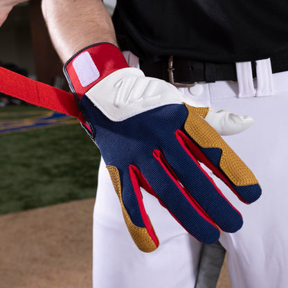 A person in a black shirt and white pants fastens the strap of Marucci Blacksmith Adult Batting Gloves (MBGBKSMFW)—featuring a blue, white, red, and tan design with a goatskin palm—while standing confidently on a sports field.