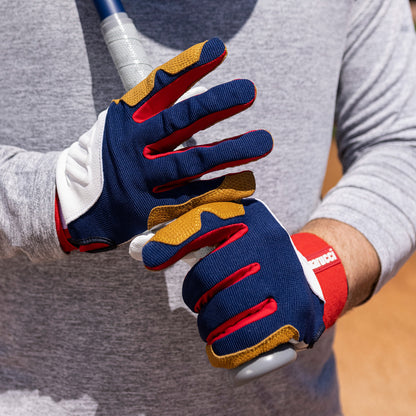 Wearing Marucci Blacksmith Adult Batting Gloves (MBGBKSMFW) in blue, red, white, and yellow with a goatskin palm, the user grips their bat firmly. The gloves pair well with a light gray long-sleeve shirt for durable batting style.