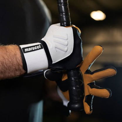 Close-up of a person wearing Marucci Blacksmith Adult Batting Gloves (MBGBKSMFW) with a durable goatskin palm, gripping a black baseball bat inside an indoor sports facility.