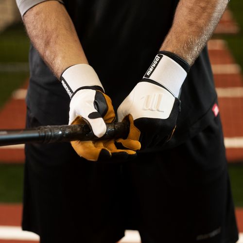 Wearing black shorts and Marucci Blacksmith Adult Batting Gloves (MBGBKSMFW), a person grips a baseball bat, ready to swing on a field with a blurry red and white background.