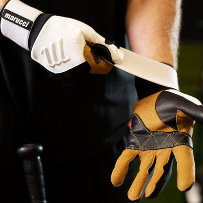 A person wearing Marucci Blacksmith Adult Batting Gloves (MBGBKSMFW) with a goatskin palm tightens the wrist strap, while a baseball bat handle appears in the foreground.