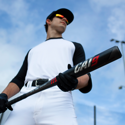 A baseball player in a black and white uniform and sunglasses holds a used 2021 Marucci CAT9 (-8) 2 3/4" USSSA Baseball Bat (MSBC98), standing outdoors with a blue sky in the background.