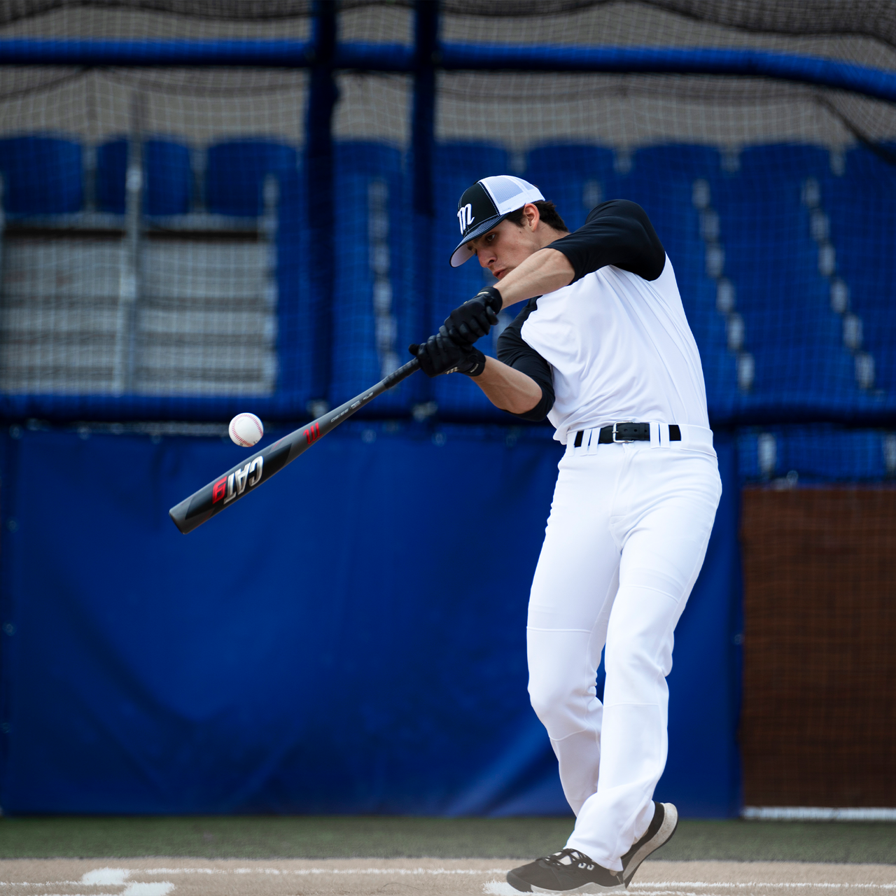 A baseball player in a white uniform with black sleeves swings a Marucci 2021 CAT9 (-8) 2 3/4" USSSA Baseball Bat (USED) to hit a pitch, with empty blue stadium seats visible in the background.