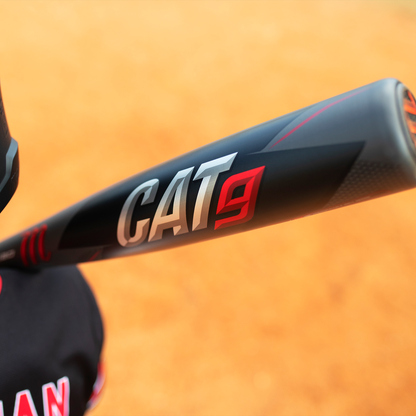 A close-up of a Marucci 2021 CAT9 (-8) 2 3/4" USSSA Baseball Bat: MSBC98 (USED), black with silver and red lettering, held by someone in a baseball uniform on a dirt field.