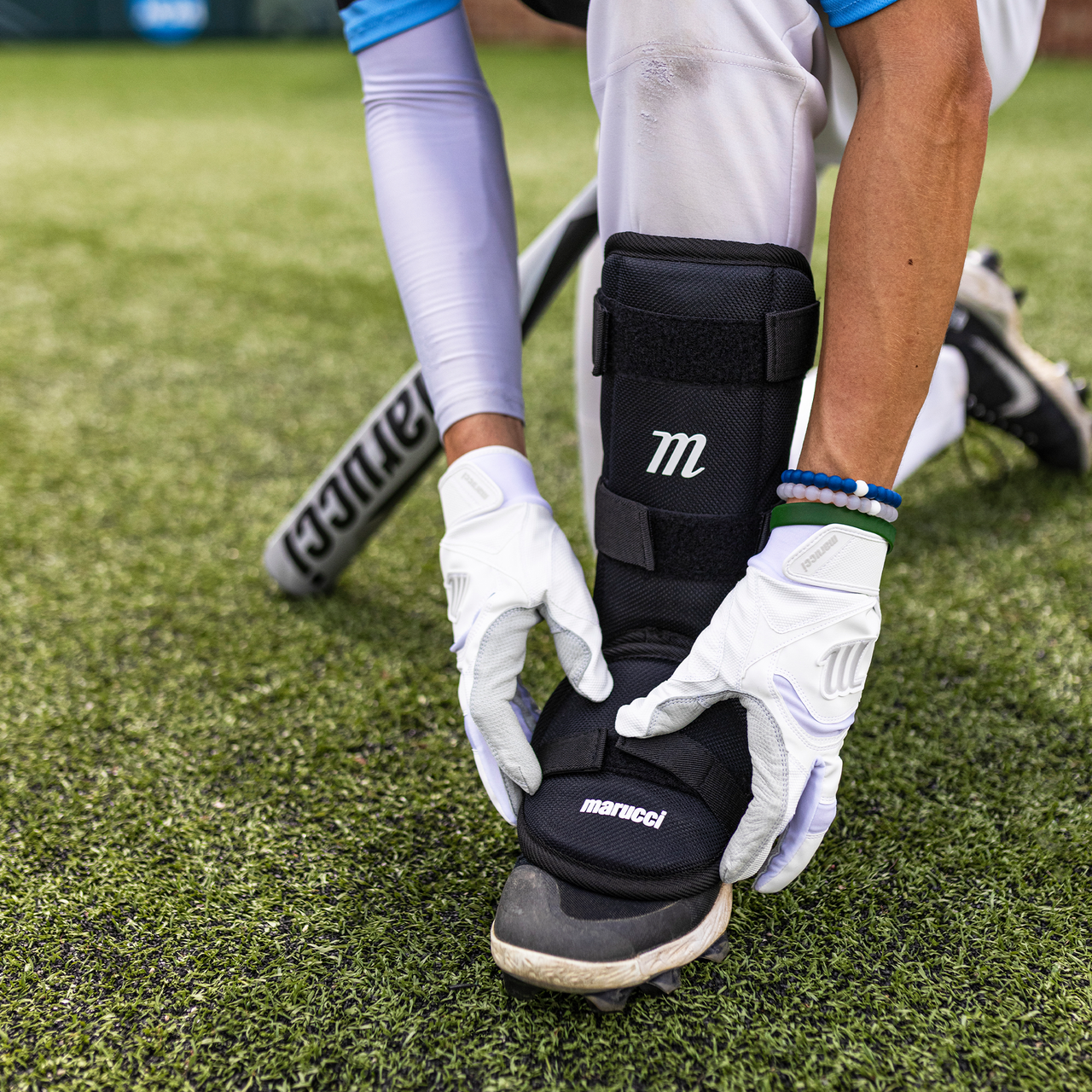 A baseball player wearing white gloves adjusts a Marucci Batter's Leg Guard (MPLG3) with extended ankle protection over their cleat on artificial turf, as a Marucci bat rests nearby.