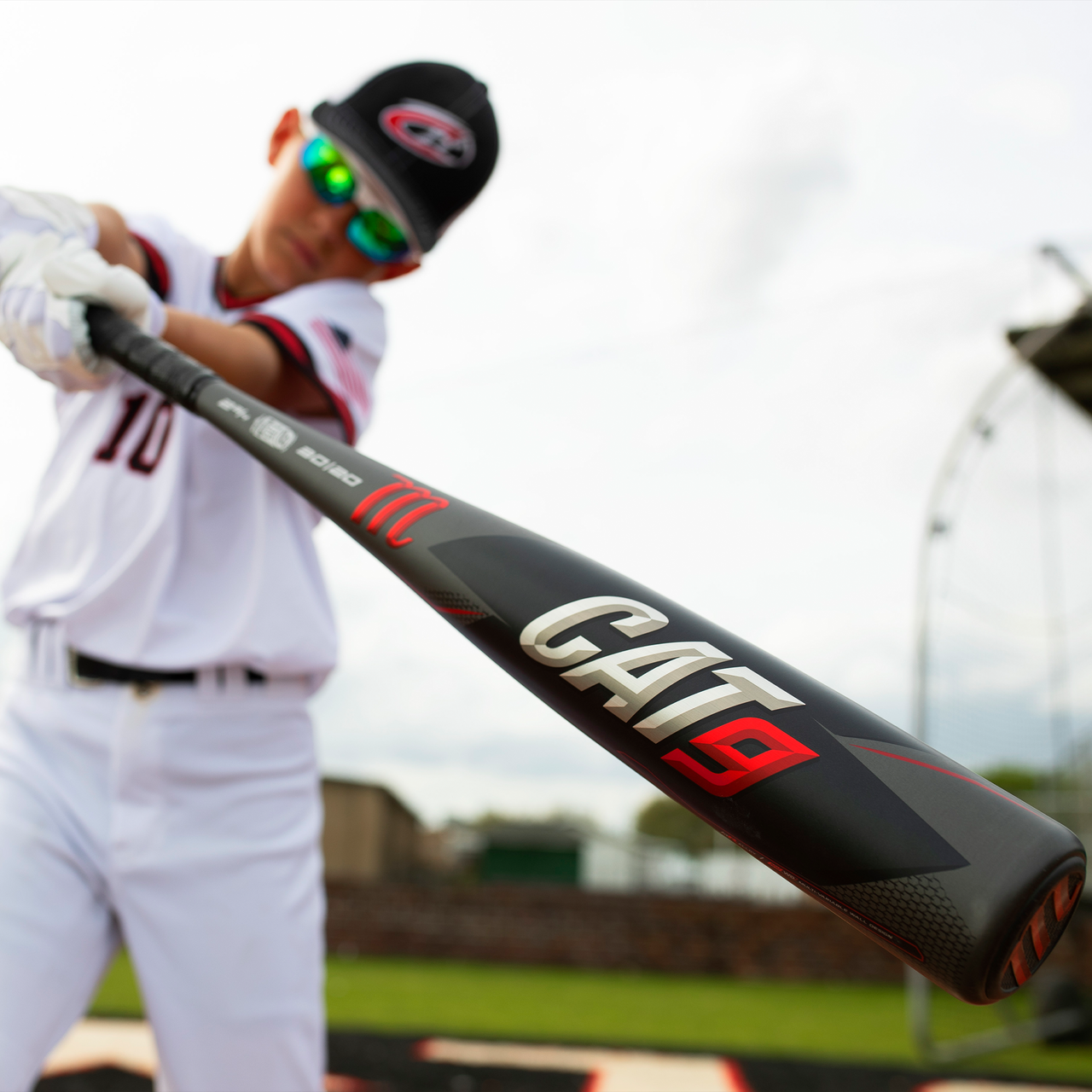 A young baseball player in uniform and sunglasses holds a black 2021 Marucci CAT9 (-8) 2 3/4" USSSA Baseball Bat: MSBC98 (USED) towards the camera, with a blurred field and batting cage in the background.