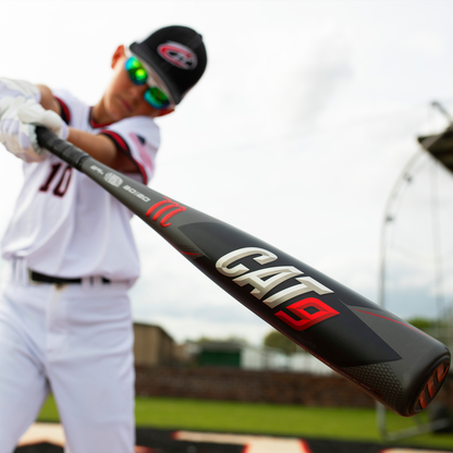 A young baseball player in uniform and sunglasses holds a black 2021 Marucci CAT9 (-8) 2 3/4" USSSA Baseball Bat: MSBC98 (USED) towards the camera, with a blurred field and batting cage in the background.