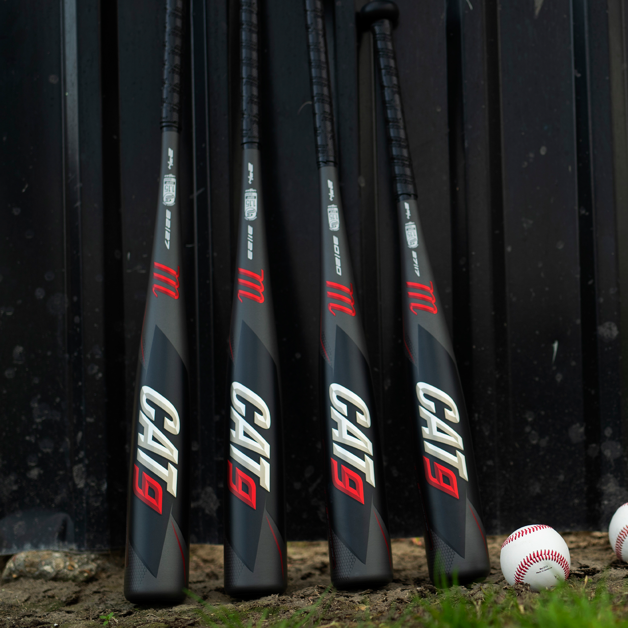 Four Marucci 2021 CAT9 (-8) 2 3/4" USSSA baseball bats (USED) stand upright against a dark wall, with three baseballs resting on the dirt nearby.
