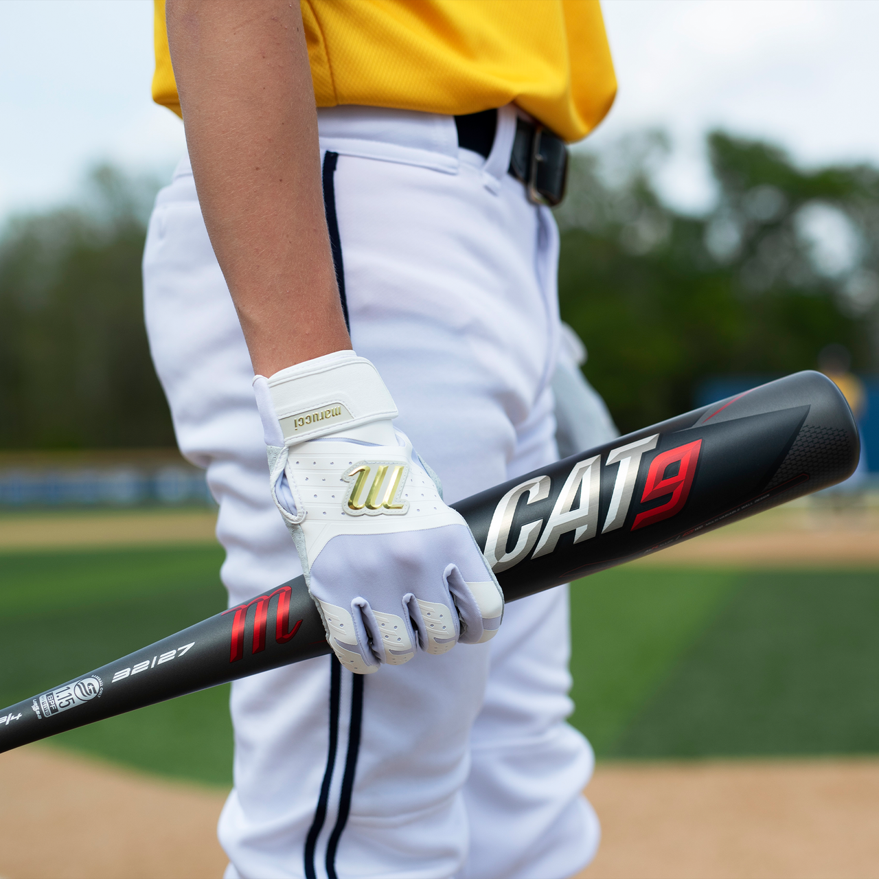 A baseball player in a yellow jersey and white pants holds a black 2021 Marucci CAT9 (-8) 2 3/4" USSSA Baseball Bat: MSBC98 (USED) and wears a white glove while standing on a baseball field.