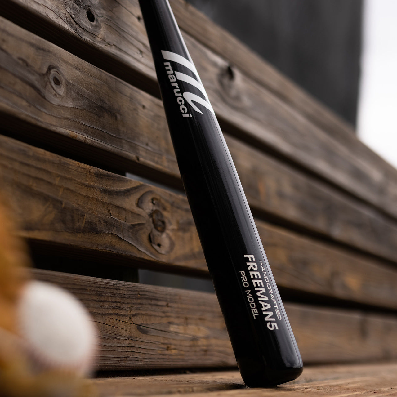 A Marucci FREEMAN5 Freddie Freeman Pro Model Maple Wood Bat (MVE2FREEMAN5-BK), an end-loaded black maple bat, sits on a wooden bench with part of a baseball nearby.