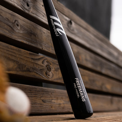 A Marucci FREEMAN5 Freddie Freeman Pro Model Maple Wood Bat (MVE2FREEMAN5-BK), an end-loaded black maple bat, sits on a wooden bench with part of a baseball nearby.
