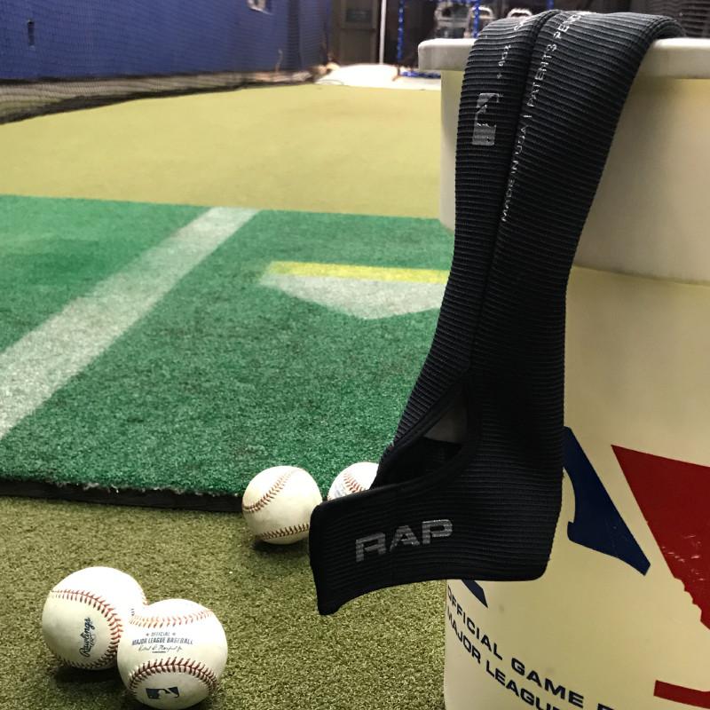 A Varo RAP Weighted Hittable Training Sleeve hangs on a large MLB baseball bucket, with several baseballs placed on artificial turf in an indoor swing speed training area.