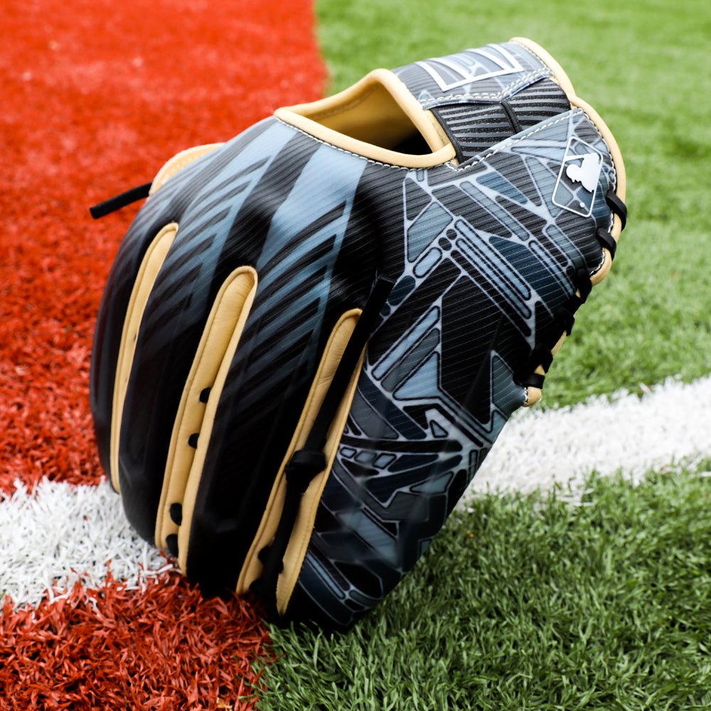 A Rawlings REV1X 12.75" Baseball Glove (REV3039-6) by Rawlings, featuring black leather, light tan laces, and abstract grey patterns, sits on artificial turf near a white line and a red section.