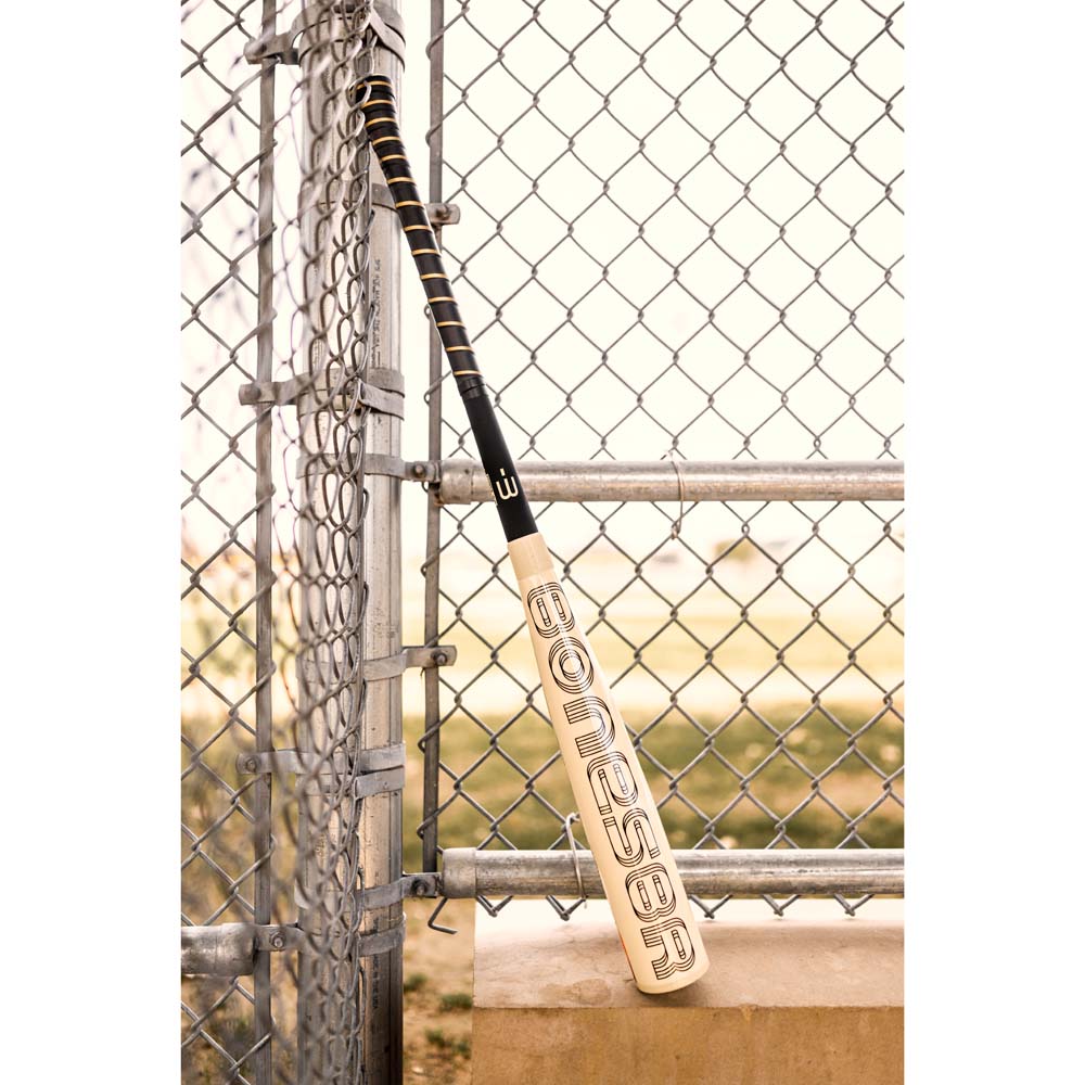 A 2024 Warstic Bonesaber Hybrid (-3) BBCOR Baseball Bat (MBBSRHB23WH3), beige in color, leans against a chain-link fence near a baseball field, with metal railing and a concrete ledge in view.