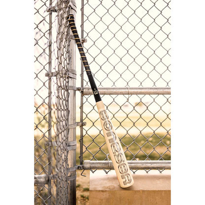 A 2024 Warstic Bonesaber Hybrid (-3) BBCOR Baseball Bat (MBBSRHB23WH3), beige in color, leans against a chain-link fence near a baseball field, with metal railing and a concrete ledge in view.