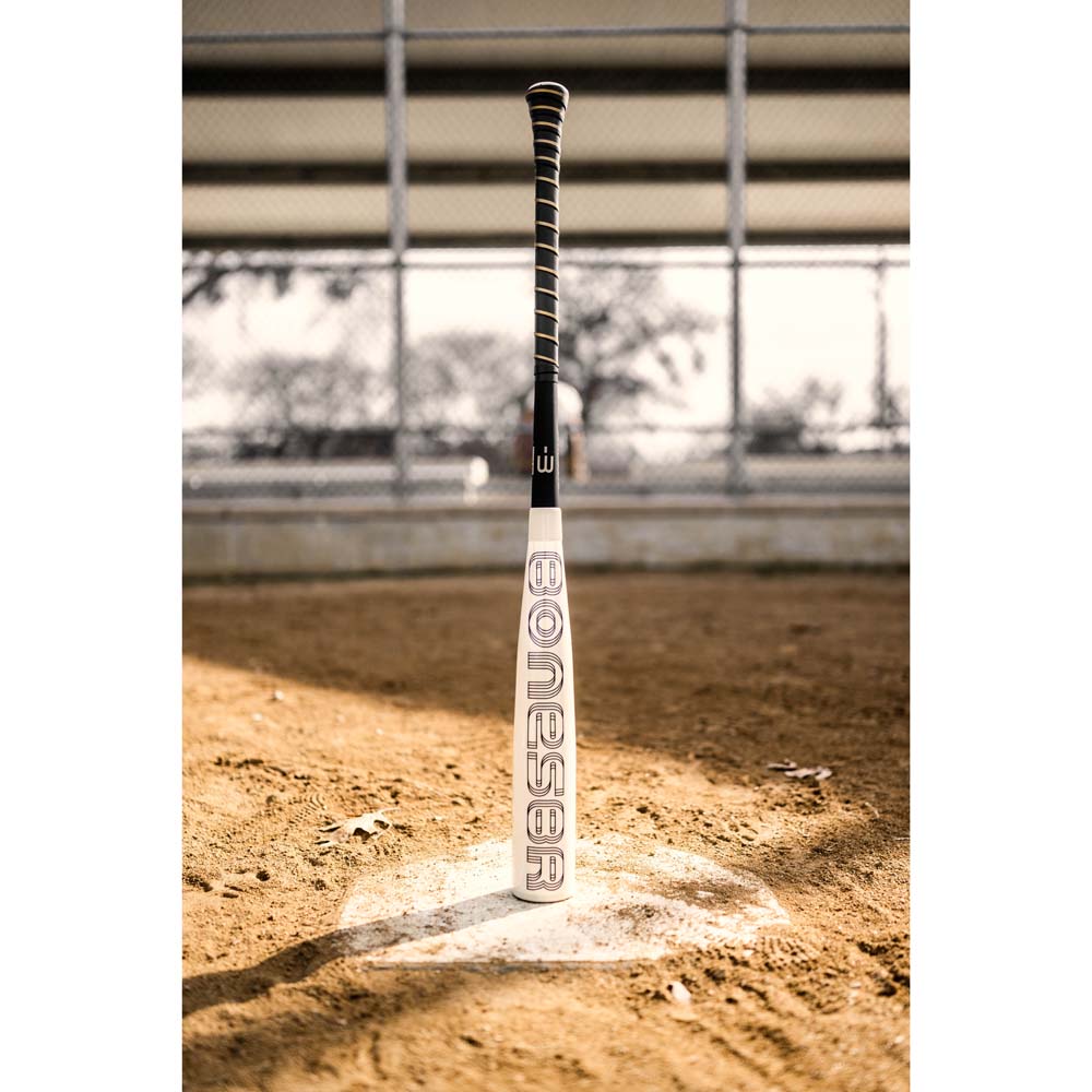 A 2024 Warstic Bonesaber Hybrid (-3) BBCOR Baseball Bat stands upright on home plate of a dirt field, with a chain-link fence and blurred background. The word "BONESABR" appears on the bat. Brand: Warstic.