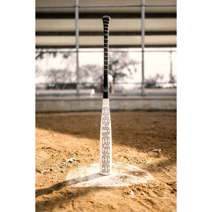 A 2024 Warstic Bonesaber Hybrid (-3) BBCOR Baseball Bat stands upright on home plate of a dirt field, with a chain-link fence and blurred background. The word "BONESABR" appears on the bat. Brand: Warstic.