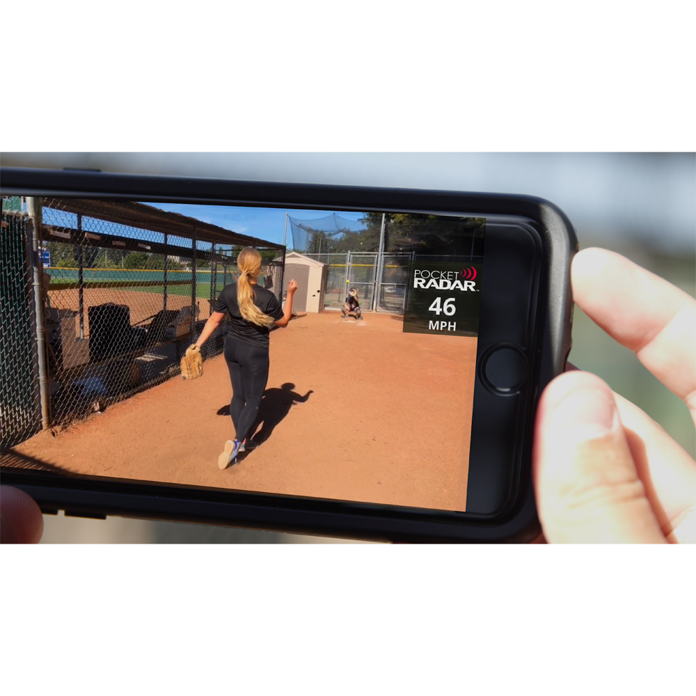 A person uses the Pocket Radar Smart Coach Radar: SR1100 by Pocket Radar, shown on a smartphone displaying a 46 MPH pitch on the app, while watching a softball pitcher throw outdoors on a sunny field.