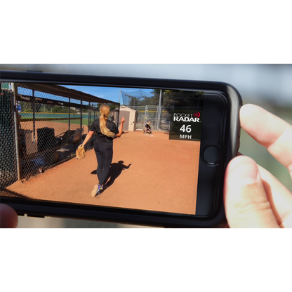 A person uses the Pocket Radar Smart Coach Radar: SR1100 by Pocket Radar, shown on a smartphone displaying a 46 MPH pitch on the app, while watching a softball pitcher throw outdoors on a sunny field.