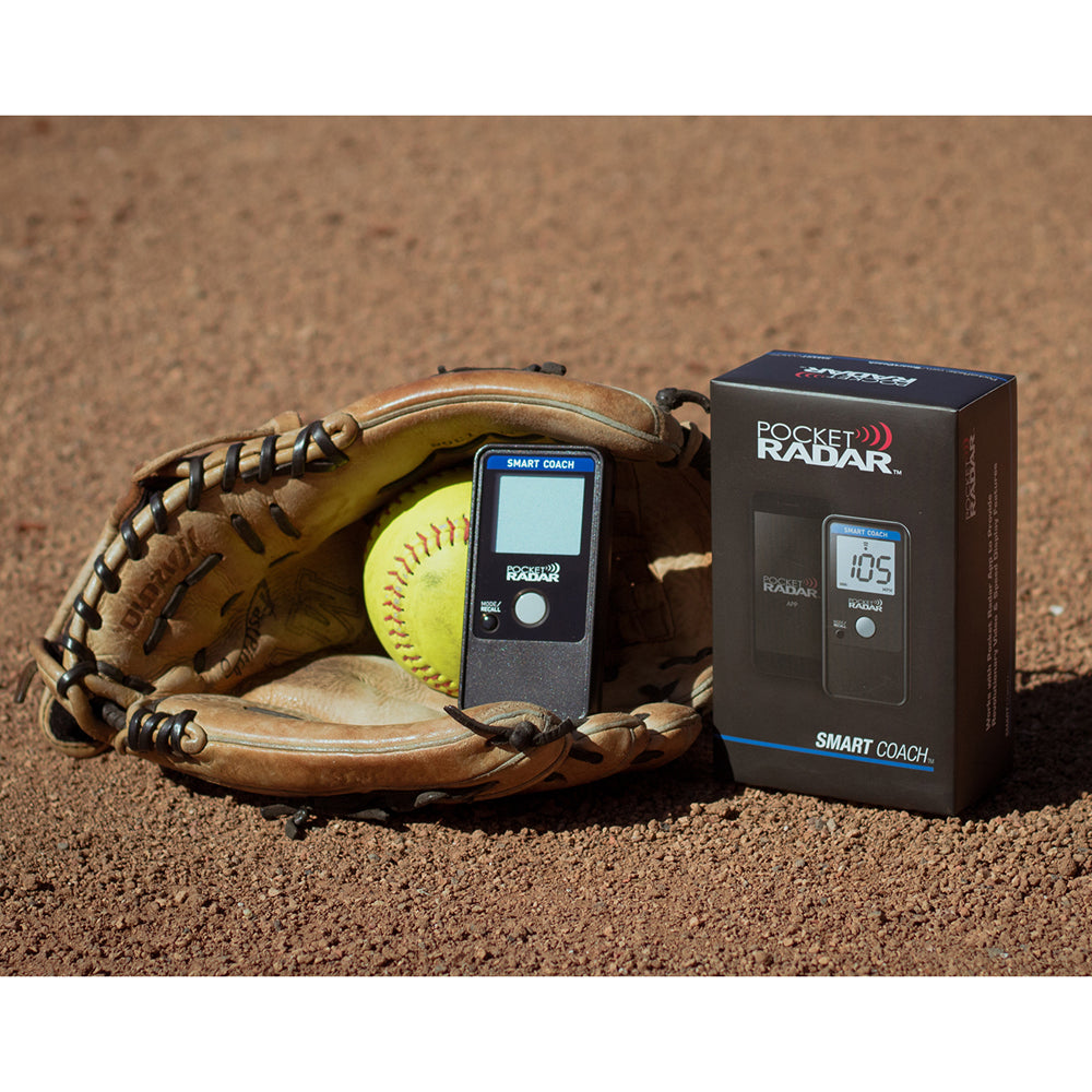 A softball glove with a yellow softball, the Pocket Radar Smart Coach Radar: SR1100 by Pocket Radar, and its box rest on a dirt field—ideal for athletes upgrading their speed training with the Pocket Radar app.