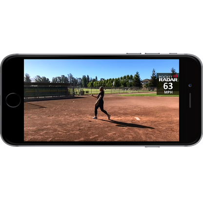 A smartphone shows a softball pitcher mid-throw on a field, with the Pocket Radar Smart Coach Radar: SR1100 by Pocket Radar app displaying a 63 MPH speed. Trees and a fence appear in the background beneath a clear sky.