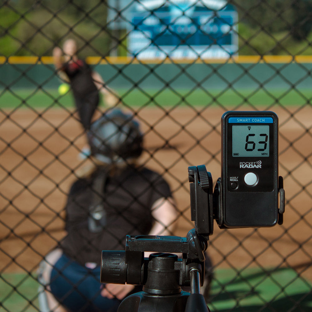 A Pocket Radar Smart Coach Radar: SR1100 by Pocket Radar, mounted on a tripod at a softball field, shows a 63 mph pitch speed while pitcher, catcher, scoreboard, and chain-link fence appear in the background.