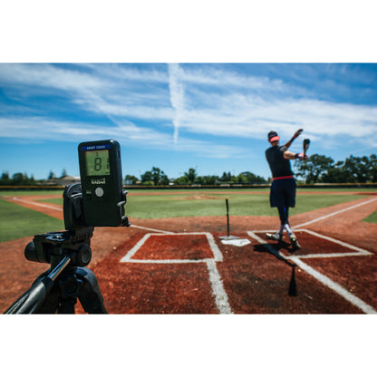 A Pocket Radar Smart Coach Radar (SR1100) by Pocket Radar, set on a tripod, measures baseball swing speed as a player hits a ball from a tee at home plate on an outdoor field under clear blue skies.